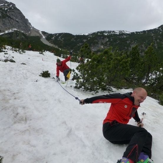 Zwei Skifahrer steigen einen schneebedeckten Berg hinauf, einer zieht den anderen mit einem Seil. Die Kulisse besteht aus schneebedeckten Bergen und grünen Bäumen.