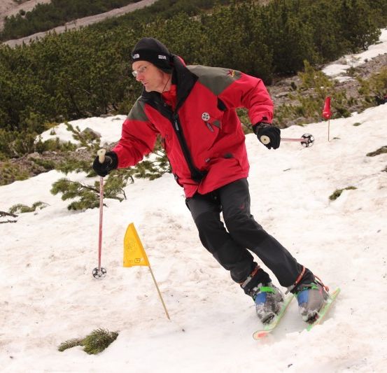 Ein Skifahrer navigiert einen verschneiten Hang, trägt einen roten Mantel, schwarze Handschuhe und Skibrille. Er hält einen Skistock und passiert eine gelbe Flagge.