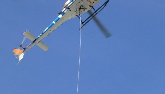 Ein Hubschrauber mit der Aufschrift 'OC-AIR' an der Seite fliegt am Himmel und trägt ein Seil.