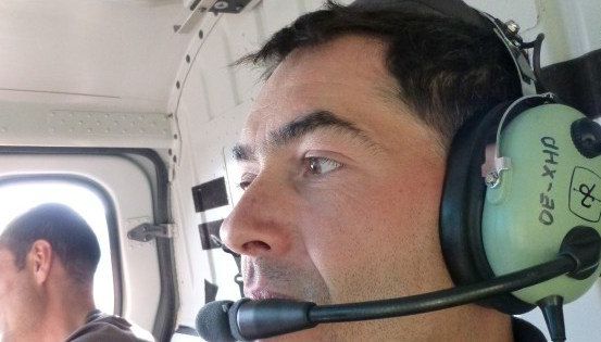 Ein Mann mit Headset und Mikrofon sitzt in einem Fahrzeug, möglicherweise einem Hubschrauber, mit einem weiteren Mann im Hintergrund.