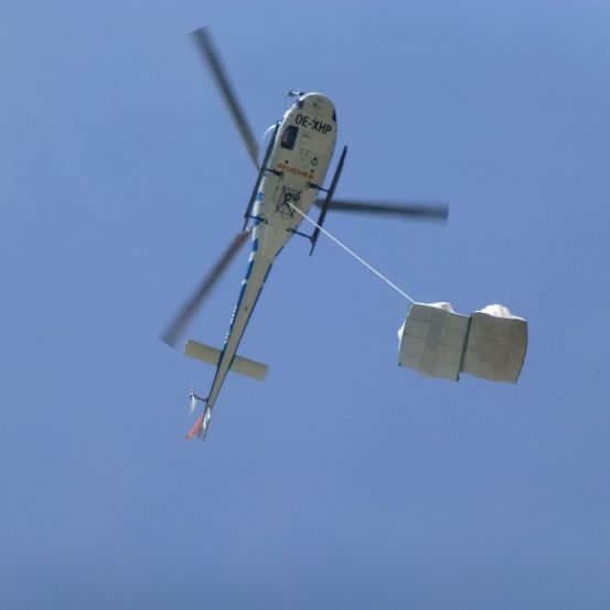 Ein Hubschrauber mit der Registrierung OE-XHP fliegt am Himmel und trägt einen Frachtcontainer an einem Seil.