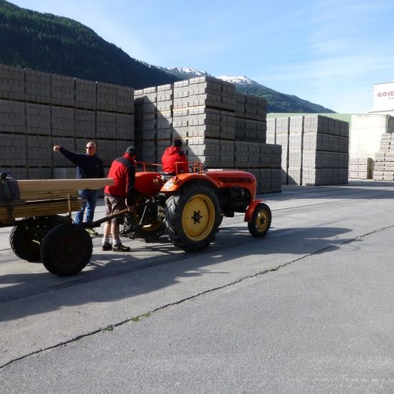 Drei Männer arbeiten mit einem roten Traktor und einem Anhänger voller Holzstämme in einem Lagerhaus.
