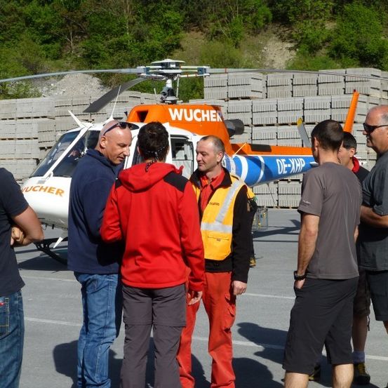 Eine Gruppe von Männern in verschiedenen farbigen Jacken steht vor einem Hubschrauber mit dem Wort Wucher darauf.