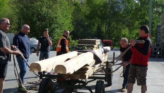 Eine Gruppe von Männern zieht einen mit Holzbrettern beladenen Karren in einem Außenbereich. Sie tragen Freizeitkleidung und scheinen zusammenzuarbeiten.