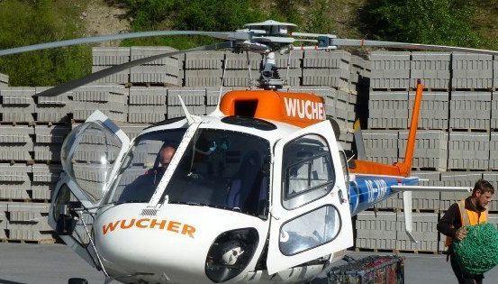 Ein Hubschrauber mit dem Wort 'WUCHER' auf der Seite ist am Boden geparkt. Ein Mann in einer gelben Weste trägt ein grünes Netz.