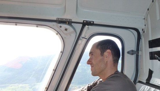 Ein Mann sitzt in einem Hubschrauber und schaut aus dem Fenster. Er lächelt und scheint die Aussicht zu genießen. Das Innere des Hubschraubers ist sichtbar, mit einem Fenster und einer Tür. Draußen gibt es Berge und einen klaren Himmel.