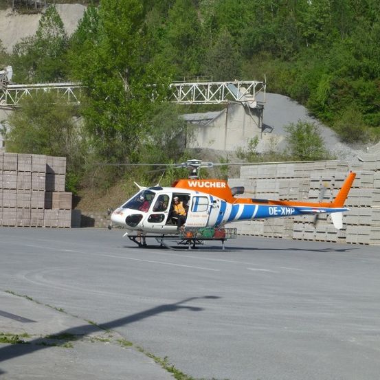 Ein Hubschrauber mit dem Kennzeichen DE-XHH steht auf einer Betonfläche in der Nähe eines Stapels Ziegel. Der Hubschrauber ist orange und weiß mit dem Wort Wucher an seiner Seite.