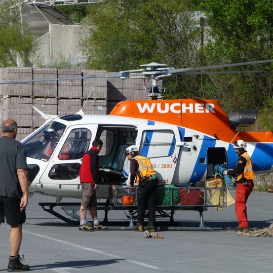 Ein Hubschrauber mit dem Wort Wucher auf der Seite ist auf dem Boden geparkt. Eine Person in einem roten Shirt steigt in den Hubschrauber ein. Eine Person in einer gelben Weste hält einen Korb.