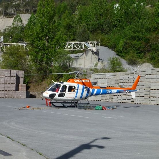 Ein orange-weißer Hubschrauber mit dem Kennzeichen DE-XHP ist auf einer Betonfläche geparkt, mit einer Person in der Nähe. Dahinter befinden sich Stapel von Ziegelsteinen und eine Metallstruktur. Bäume und ein Berg sind im Hintergrund zu sehen.