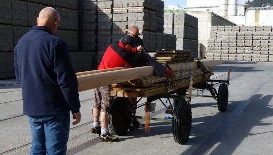 Drei Männer laden Holzbretter auf einen Karren in einem Lagerhaus. Ein Mann trägt ein rotes Shirt und einen schwarzen Hut, ein anderer ein blaues Jackett. Der Karren ist mit Holzbrettern gefüllt.