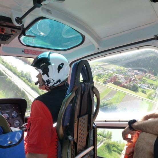 In einem Hubschrauber fliegt ein Pilot mit weißem Helm und rotem Shirt über ein grünes Tal mit Häusern und Bäumen. Ein Passagier sitzt neben ihm.