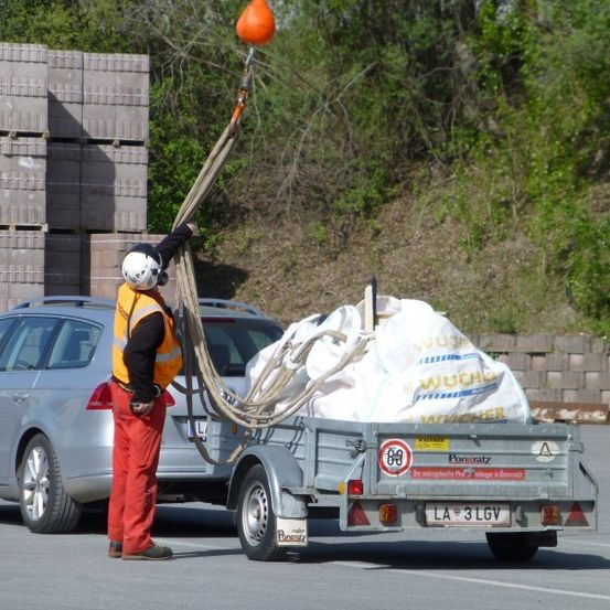 Eine Person in einer Warnweste und einem Helm benutzt einen Kran, um einen Zementsack von einem Anhänger hinter einem silbernen Auto zu heben.