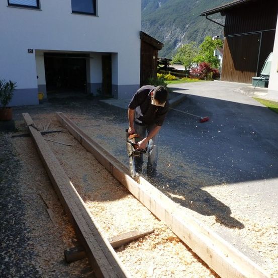 Ein Mann sägt mit einer Kettensäge einen Holzbalken auf einer Einfahrt vor einem Haus.