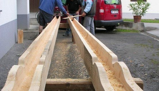 Drei Männer arbeiten an großen Holzbalken im Freien. Einer formt das Holz mit einem Werkzeug, während die anderen beiden beobachten. Ein roter Van ist in der Nähe geparkt.