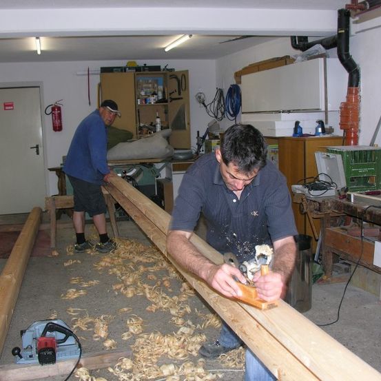 Zwei Männer arbeiten in einer Werkstatt, einer benutzt einen Hobel auf einem Holzbalken, während der andere steht und beobachtet. Der Raum ist mit Werkzeugen und Holzmaterialien gefüllt.