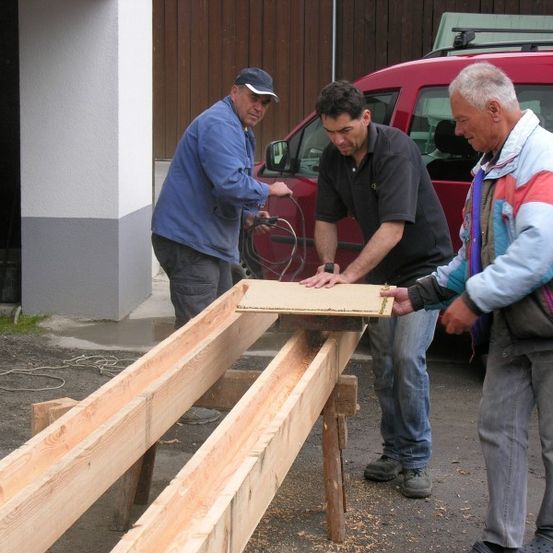 Drei Männer arbeiten im Freien an einer Holzstruktur. Ein Mann hält eine Holzplatte, während ein anderer sie betrachtet. Der dritte Mann hält ein Kabel. Ein roter Van ist hinter ihnen geparkt.