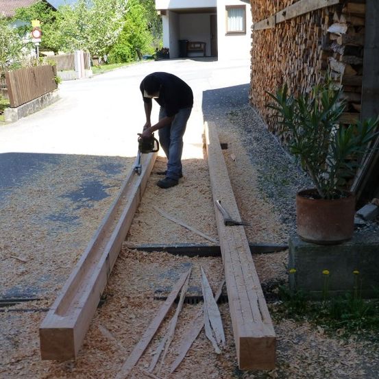 Ein Mann sägt mit einer Kettensäge einen langen Holzbalken auf einem Einfahrtsweg, neben einem Topf mit Pflanzen und einem Stapel Holz.
