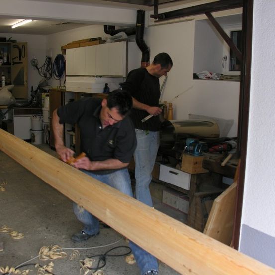 Zwei Männer arbeiten in einer Garage, einer sägt an einem langen Holzbalken, während der andere einen Messstab hält.