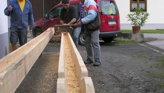 Drei Männer arbeiten an Holzbalken auf einem Parkplatz. Einer benutzt eine Säge, ein anderer hält einen Holzbalken und der dritte steht in der Nähe. Ein roter Lieferwagen ist dahinter geparkt.
