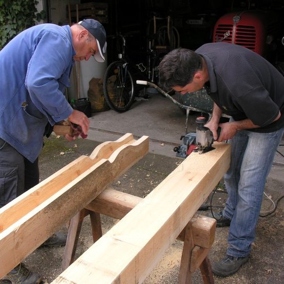 Zwei Männer arbeiten in einer Garage an Holzbalken. Einer benutzt einen Meißel, der andere eine Kreissäge. Ein roter Traktor und ein Fahrrad sind im Hintergrund.