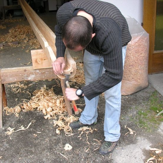 Ein Mann benutzt einen Hammer, um einen Holzbalken auf einer Werkbank zu formen, umgeben von Holzspänen.