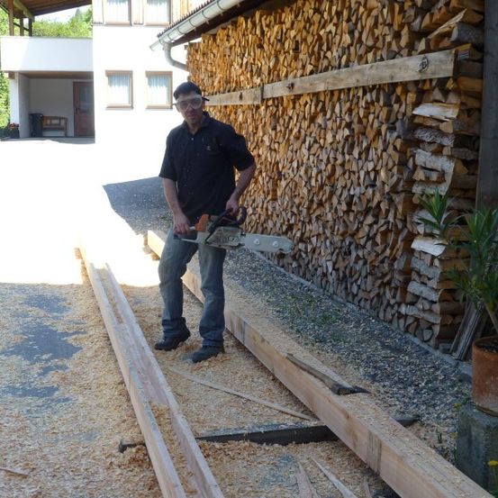 Ein Mann mit Schutzbrille und schwarzem Shirt schneidet Holz mit einer Kettensäge vor einem Haus mit einem großen Holzstapel.