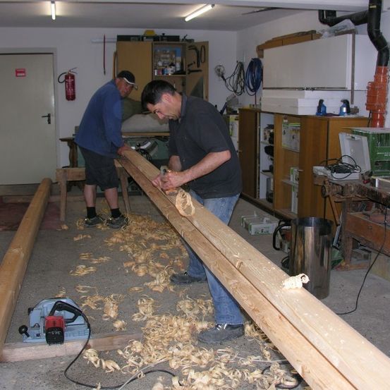 Zwei Männer arbeiten in einer Garage, einer benutzt eine Kreissäge, um einen langen Holzbalken zu schneiden, während der andere zuschaut.