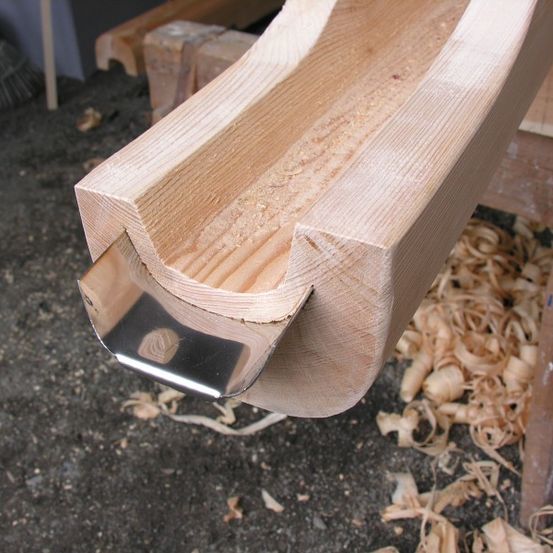 Ein Nahaufnahme einer Holzbalken mit einem Metallwerkzeug, das zeigt, wie das Holz geformt wird. Der Balken befindet sich auf einer Werkbank mit verstreutem Sägemehl.