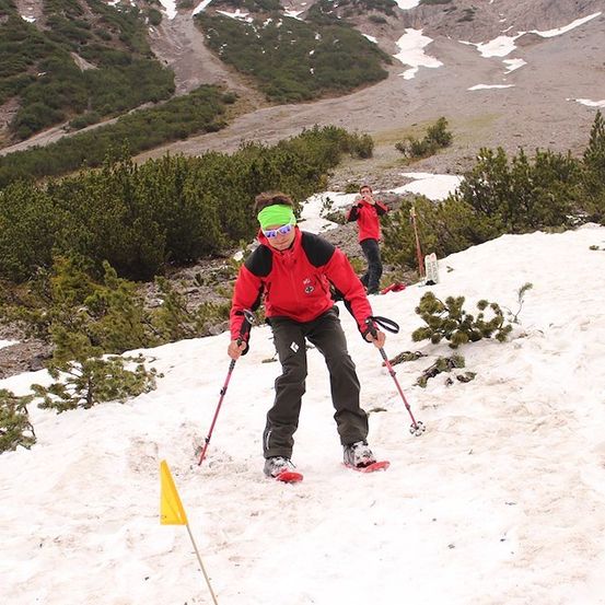 Ein Skifahrer in einem roten Jackett und einem grünen Stirnband navigiert einen verschneiten Berg mit Skistöcken. Hinter ihm macht ein weiterer Skifahrer in einem roten Jackett ein Foto.