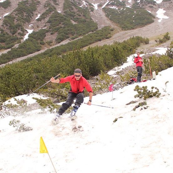 Ein Skifahrer in einem roten Jackett und schwarzen Hosen fährt einen schneebedeckten Berg hinunter und hält Skistöcke. Ein weiterer Skifahrer in Rot steht im Hintergrund.