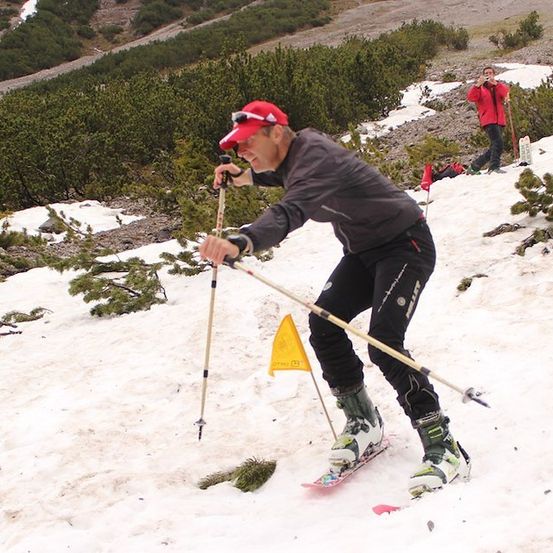 Ein Mann skifahrer auf einem verschneiten Berg, hält Skistöcke und eine gelbe Flagge. Eine weitere Person ist im Hintergrund und macht ein Foto.