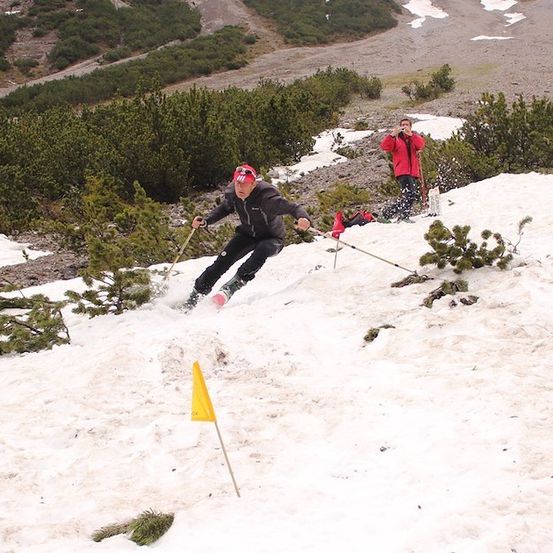 Ein Skifahrer in einem roten Jackett und schwarzen Hosen fährt einen schneebedeckten Hang hinunter mit einer gelben Fahne. Ein weiterer Skifahrer in einem roten Jackett steht hinter ihm.