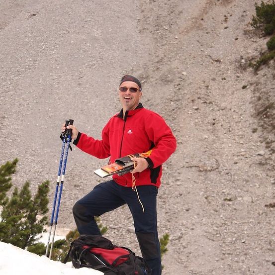 Ein Mann in einem roten Jackett und blauen Hosen steht auf einem verschneiten Hang, hält Skistöcke und eine Karte. Er lächelt und scheint die Aussicht zu genießen.