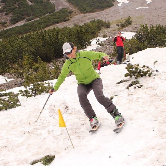 Ein Skifahrer in einer grünen Jacke und grauen Hosen fährt einen schneebedeckten Hang hinunter und hält Skistöcke. Ein gelber Flaggen markiert den Weg. Im Hintergrund beobachtet ein anderer Skifahrer in einer roten Jacke.