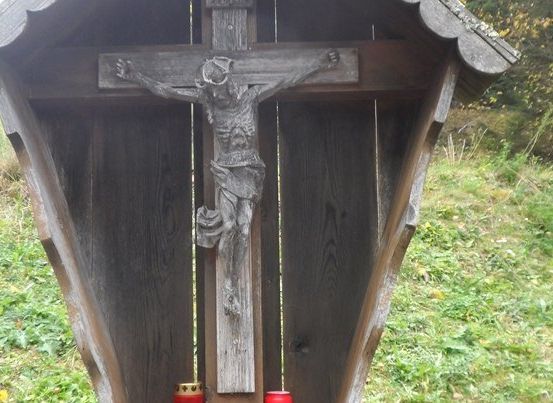 Ein hölzernes Kruzifix mit einer Statue von Jesus Christus am Kreuz, in einer kleinen Holzkapelle mit zwei roten Kerzen auf beiden Seiten platziert.