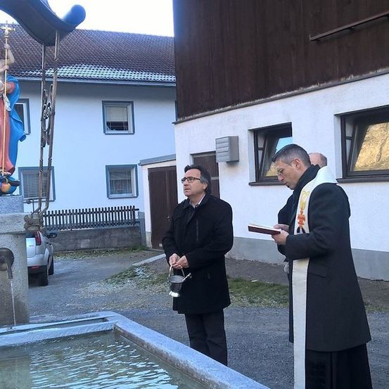 Zwei Männer stehen neben einem Brunnen mit einer Statue von Maria und Jesus, einer hält ein Buch und der andere einen Eimer.