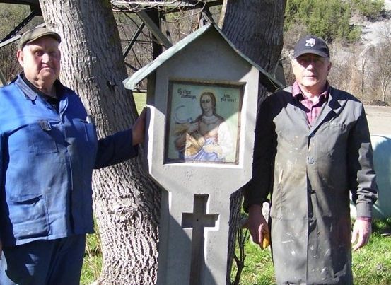 Zwei Männer stehen neben einem Betonkreuz mit einem religiösen Bildnis, einer hält ein Werkzeug und der andere einen Pinsel.
