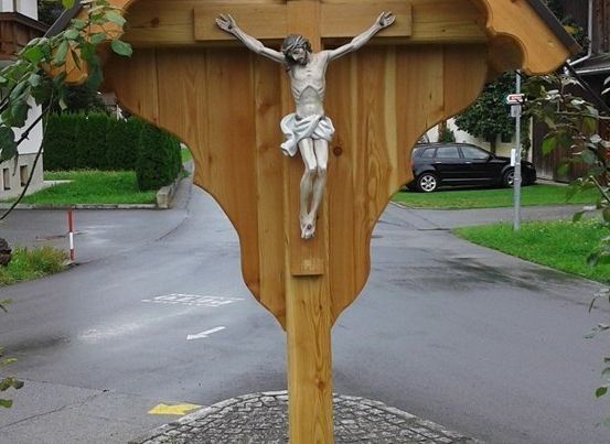 Ein hölzernes Kruzifix steht an einer Straßenecke, umgeben von Grünpflanzen und einem kleinen Kreisverkehr. Ein schwarzes Auto ist in der Nähe geparkt.