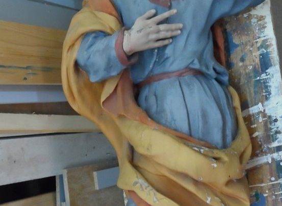 Eine Statue einer Person in blauen und gelben Gewändern wird repariert. Die Statue lehnt sich gegen eine Holzkonstruktion mit einer weißen Wand im Hintergrund.