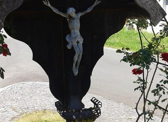 Ein Kruzifix, das an einer Holzstruktur befestigt ist, umgeben von roten Rosen, mit einem Grasbereich und einem Gebäude im Hintergrund.