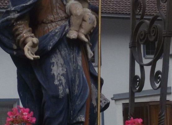 Eine Statue einer Frau, die ein Kind hält, gekleidet in Blau, steht vor einem weißen Gebäude mit einem roten Dach.