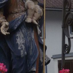 Eine Statue einer Frau, die ein Kind hält, gekleidet in Blau, steht vor einem weißen Gebäude mit einem roten Dach.