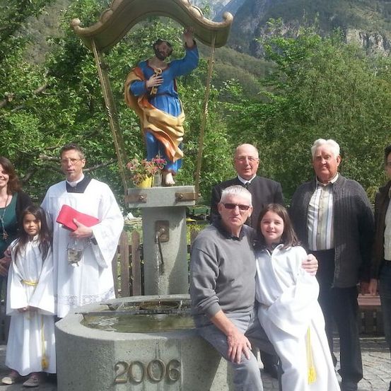 Eine Gruppe von Menschen, einschließlich Kinder, posiert für ein Foto vor einer Statue und einem Brunnen. Der Mann mit den Brillen sitzt auf einer Bank.
