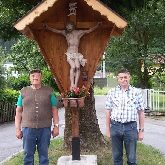 Zwei Männer stehen neben einem hölzernen Kruzifix mit einer Statue von Jesus Christus am Kreuz, umgeben von Blumen und Grün.