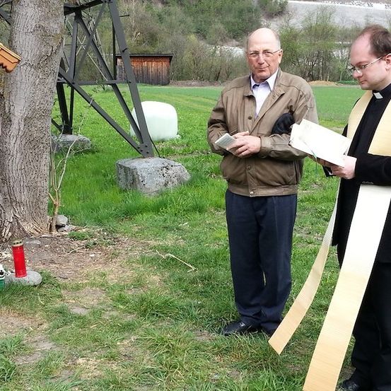Zwei Männer, einer in einer braunen Jacke und der andere in einem schwarzen und gelben Gewand, stehen auf einem Feld und lesen aus Büchern. Ein kleines Heiligtum mit einem Kreuz ist in der Nähe.