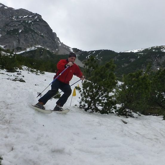 Ein Skifahrer in einem roten Jackett fährt einen schneebedeckten Berghang hinunter und hält Skistöcke.