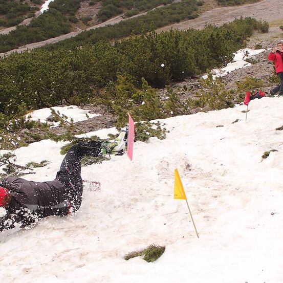 Ein Snowboarder stürzt auf einem verschneiten Berg, mit einer gelben Fahne und einer weiteren Person im Hintergrund.