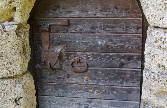 Eine alte Holztür mit einem verrosteten Metallschloss und einem Schlüsselloch, in eine Steinmauer eingelassen.