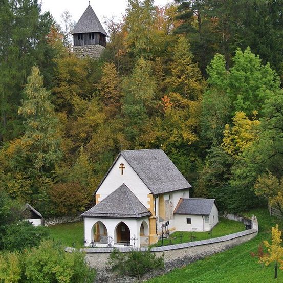 Eine kleine Kirche mit grauem Dach und Turm ist von Bäumen und Gras umgeben.