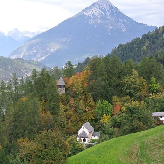 Ein malerisches Dorf in den Bergen, mit einem Turm und einer Kapelle, umgeben von herbstlich gefärbten Bäumen.